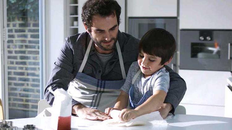 A dad helps his son as they roll out some dough.